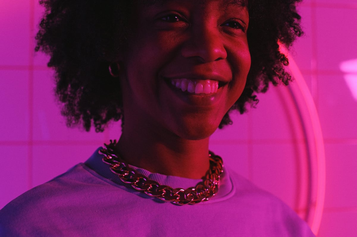 Smiling person with curly hair wearing a purple shirt and gold chain necklace under vibrant pink neon lighting